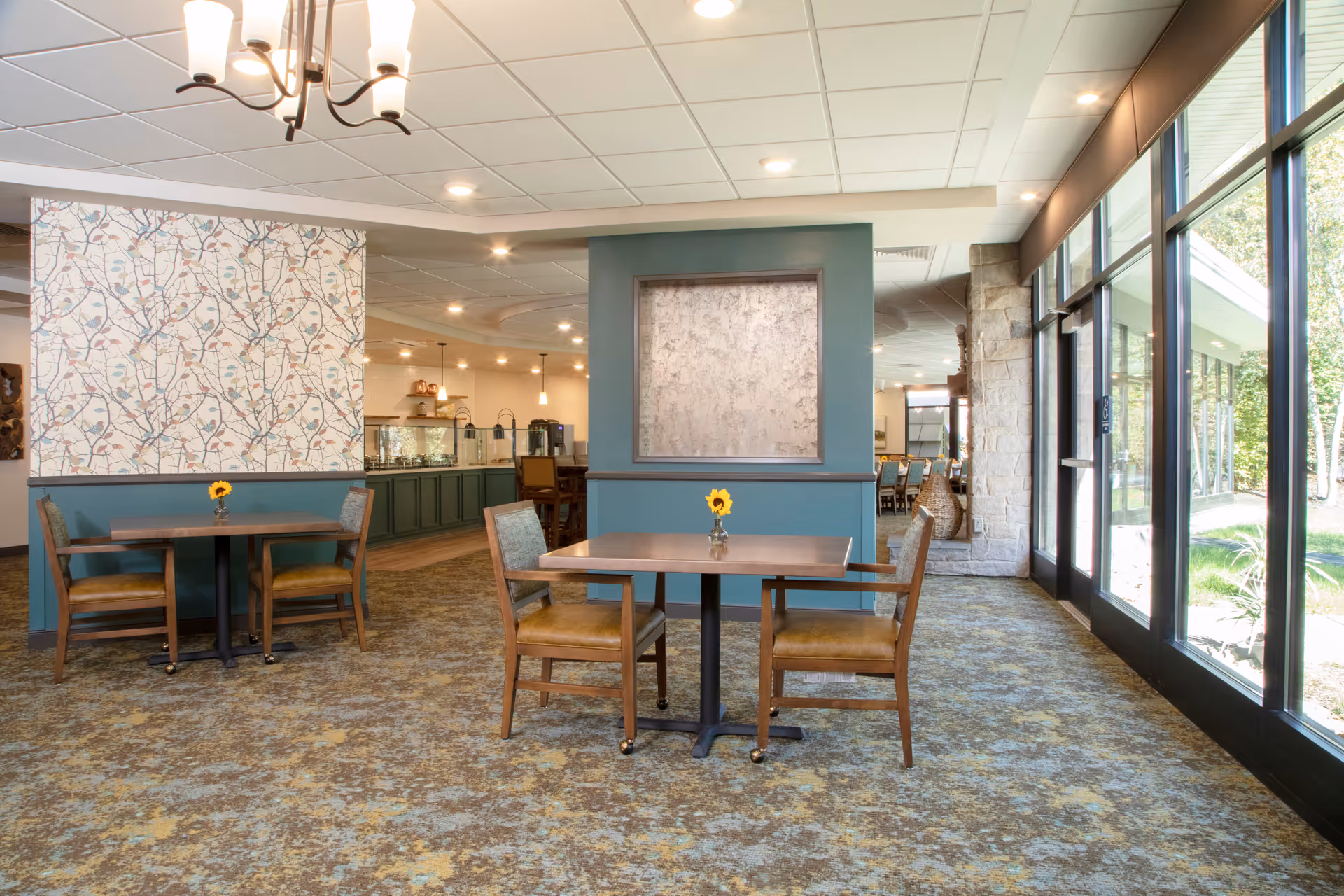 A bright dining area with two wooden tables, each accompanied by two wooden chairs with cushioned seats. Each table has a small vase with a single yellow flower. The room features large windows on the right side letting in natural light and offering a view of greenery outside. The walls are decorated with patterned wallpaper and a large framed textured panel. The floor is carpeted with a patterned design, and there is a chandelier hanging from the ceiling.