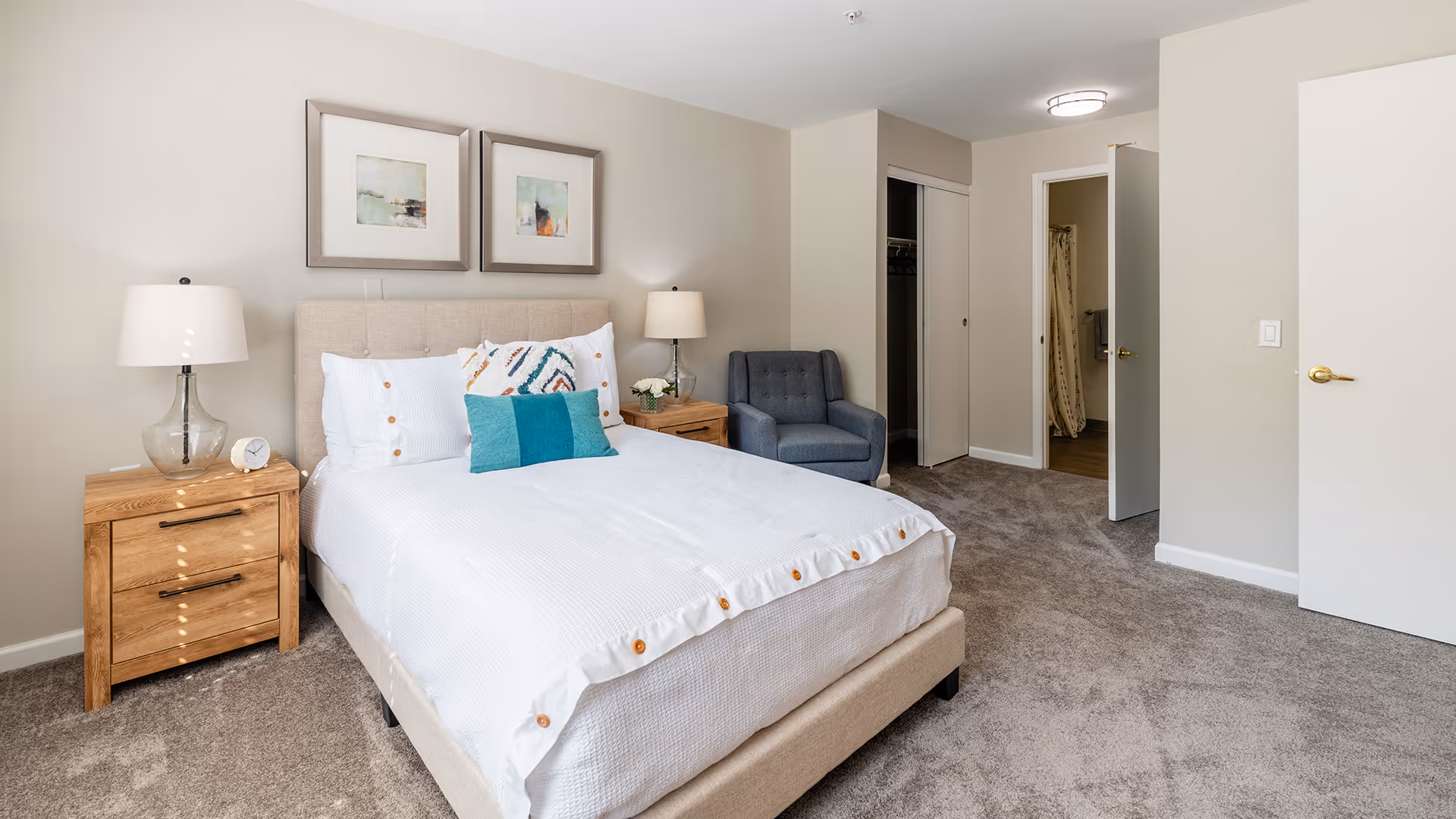 A neatly made bed with white bedding and decorative pillows in a bedroom. There are two wooden nightstands with lamps on either side of the bed. A blue armchair is placed in the corner near an open door leading to a bathroom. The room has beige walls and carpeted flooring.