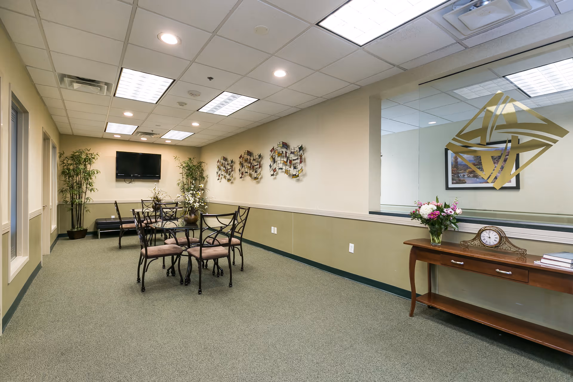 A well-lit interior room with a green carpet and beige walls. There are two small round tables with four chairs each, a wall-mounted TV, decorative plants, and metal wall art. On the right side, a wooden console table holds a clock, a vase with flowers, and some books. A large window with a gold geometric decal looks into another room.