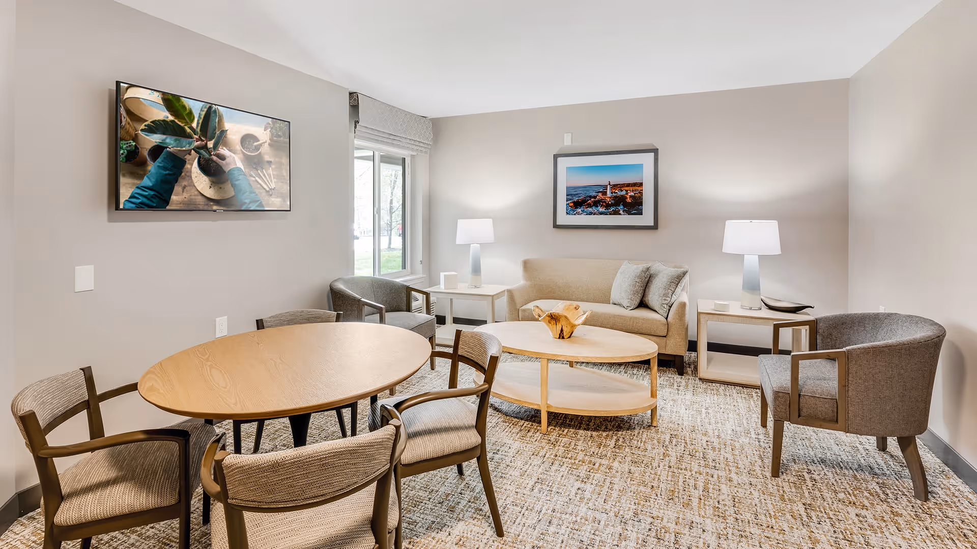 A cozy living room with a beige sofa, two gray armchairs, a wooden oval coffee table, and a round wooden dining table with four chairs. The room has neutral-colored walls, a carpeted floor, two table lamps on side tables, a large window with a view outside, and two framed pictures on the walls.