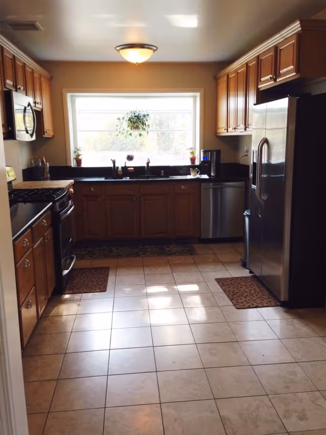Kitchen with tiled floor, wooden cabinets, stainless steel refrigerator and stove, and a large window above the sink.