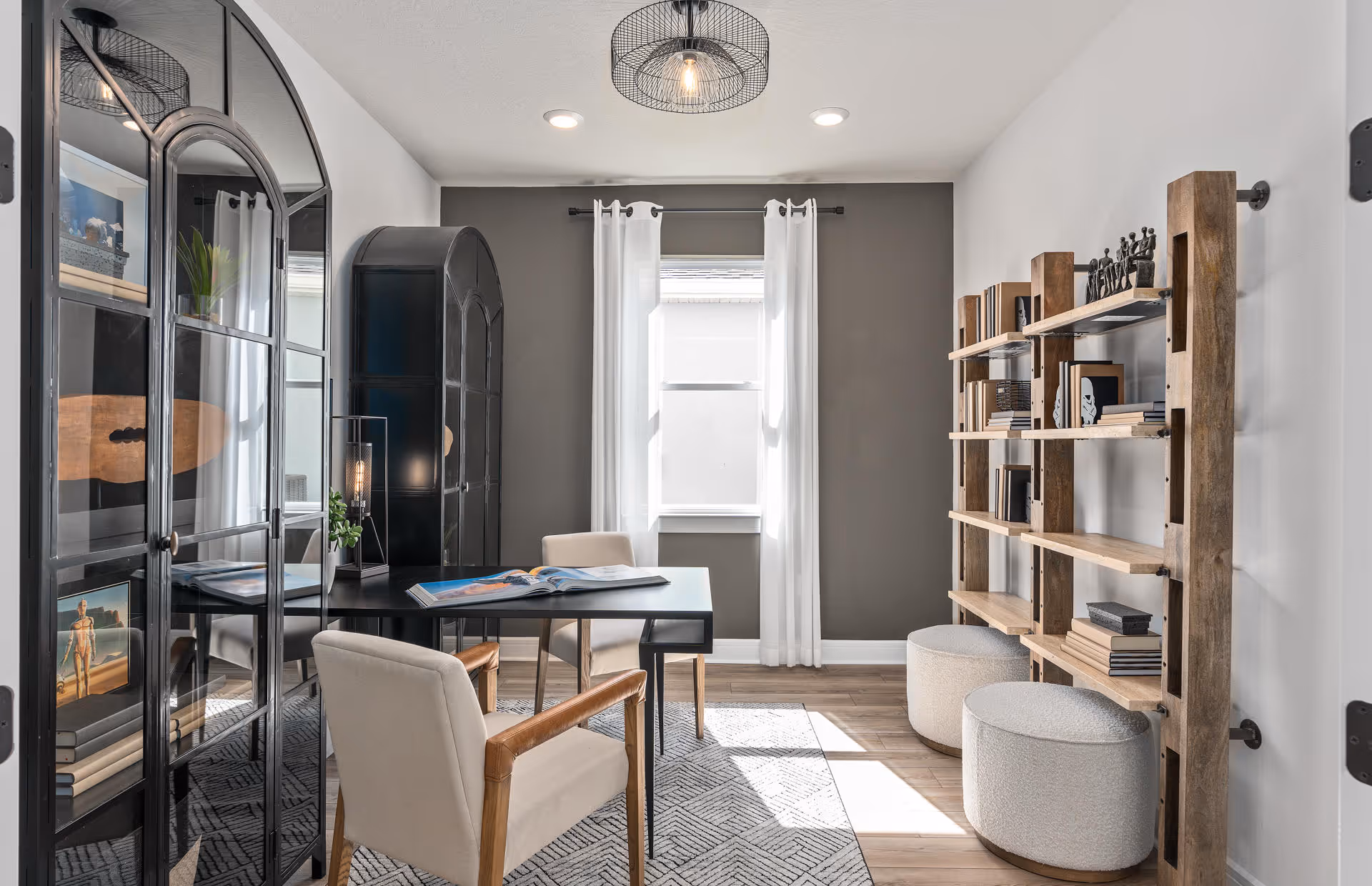 Bright modern home office with a black desk, upholstered chairs, glass-front cabinets, wooden shelving and a window with white curtains.