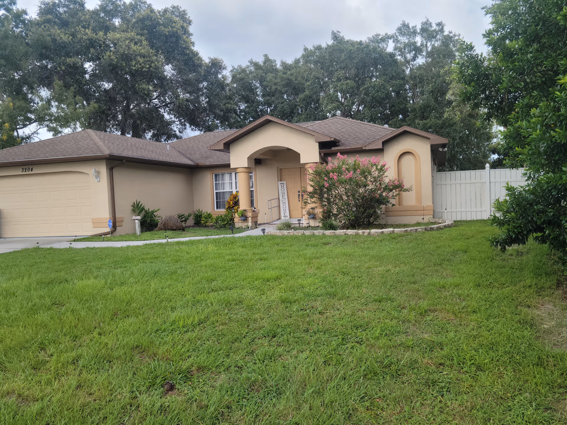 Single-story beige house with an attached garage, front lawn, and landscaped entry under a cloudy sky.