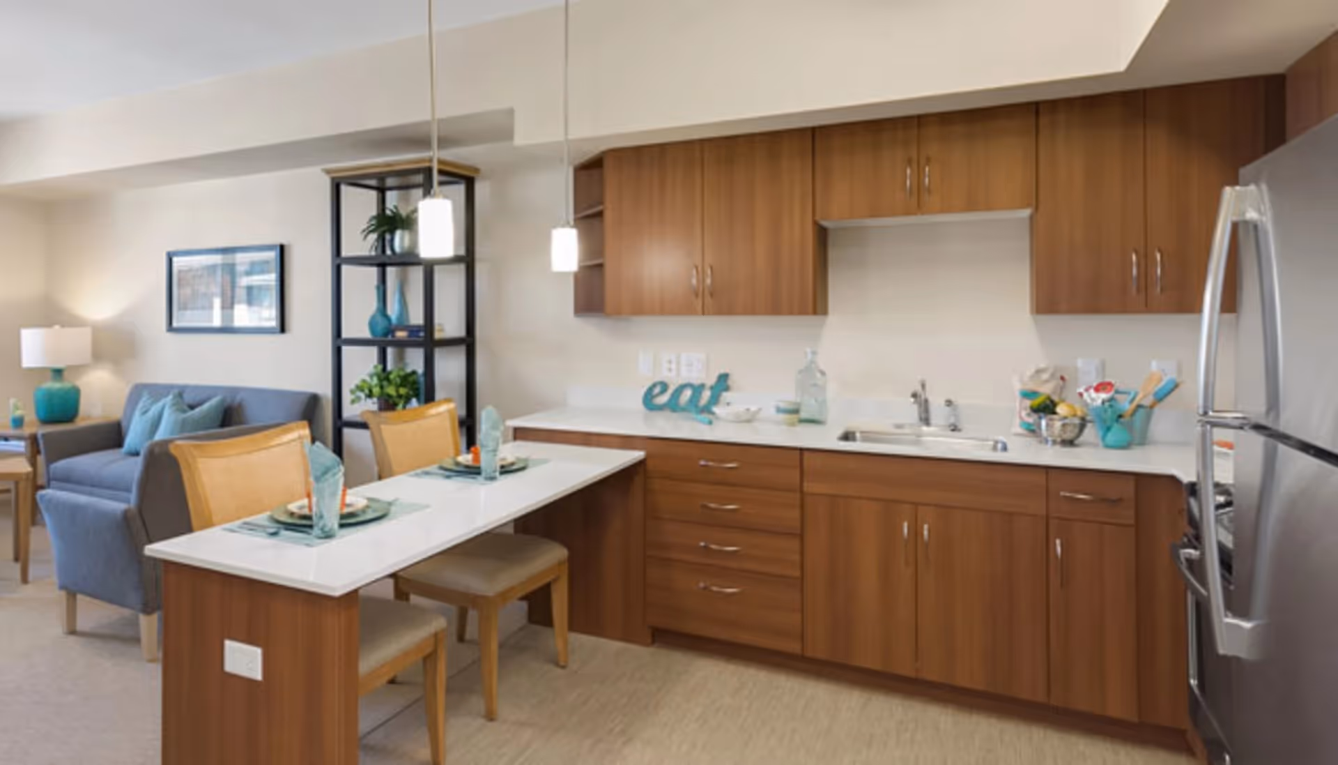 A modern kitchen and dining area in a senior living facility with wooden cabinets, a stainless steel refrigerator, a white countertop with a sink, and a small dining table set for two with chairs. In the background, there is a living room area with a gray sofa, a side table with a lamp, and a black shelving unit with decorative items.