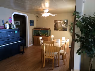 A dining area with a wooden table and six matching chairs on a wooden floor. There is a large fish tank against the wall, a ceiling fan with lights above the table, a piano on the left side, and a large plant on the right. A framed picture hangs on the wall behind the fish tank.