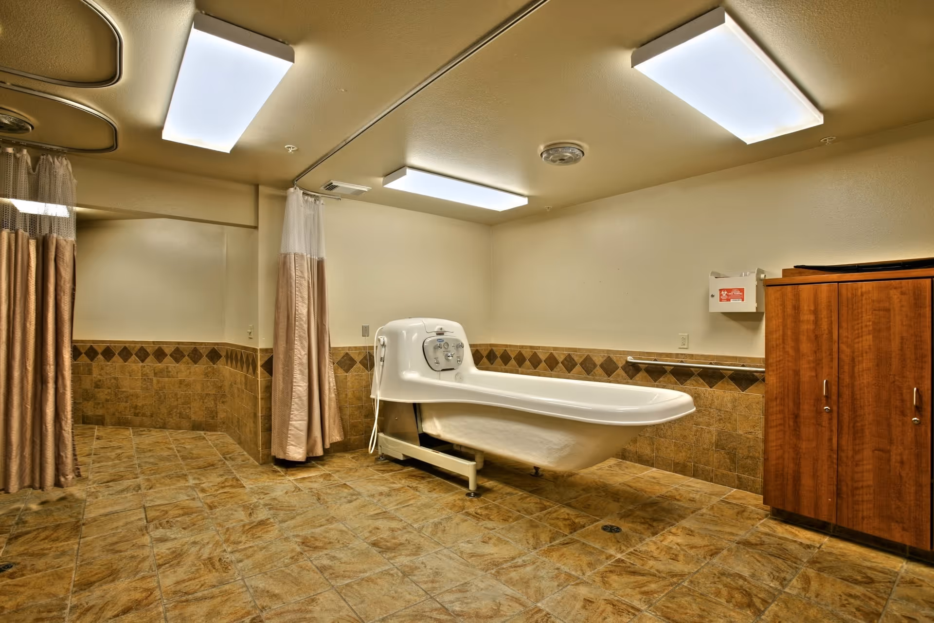 A spacious bathroom with tiled floors and walls, featuring a large white medical bath tub with controls on one end. There are beige privacy curtains hanging from ceiling tracks and a wooden cabinet on the right side. The room is well-lit with rectangular ceiling lights.
