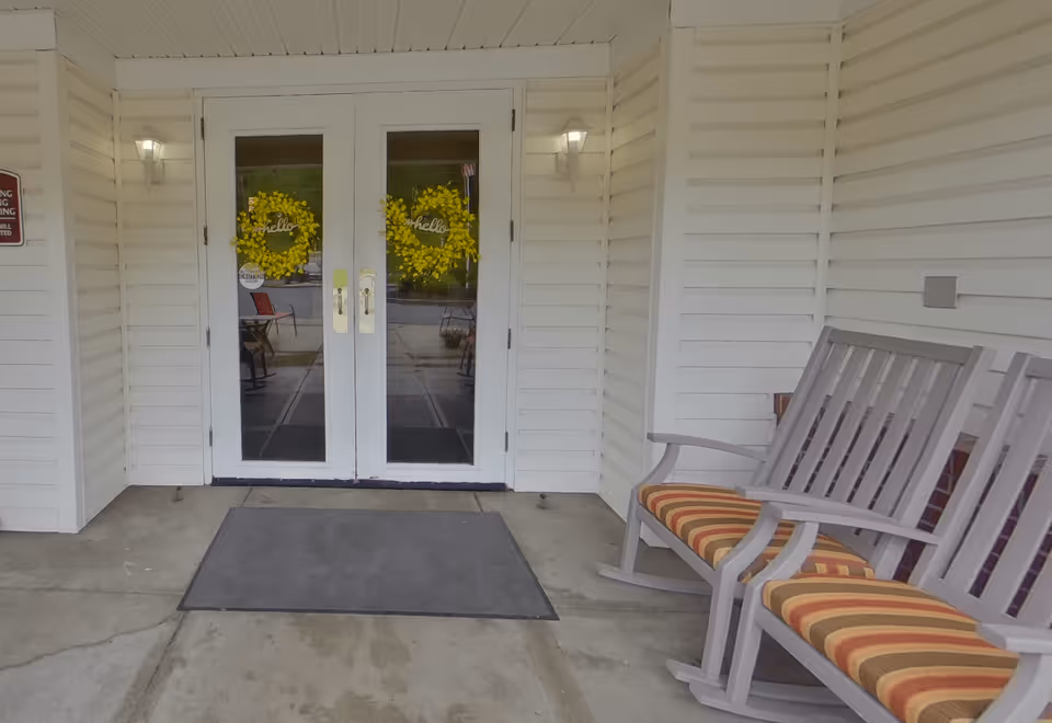 Entrance to a building with white double doors decorated with yellow wreaths that say 'hello'. There are two wall-mounted lights on either side of the doors, a gray doormat on the concrete floor, and two wooden rocking chairs with striped cushions on the right side.