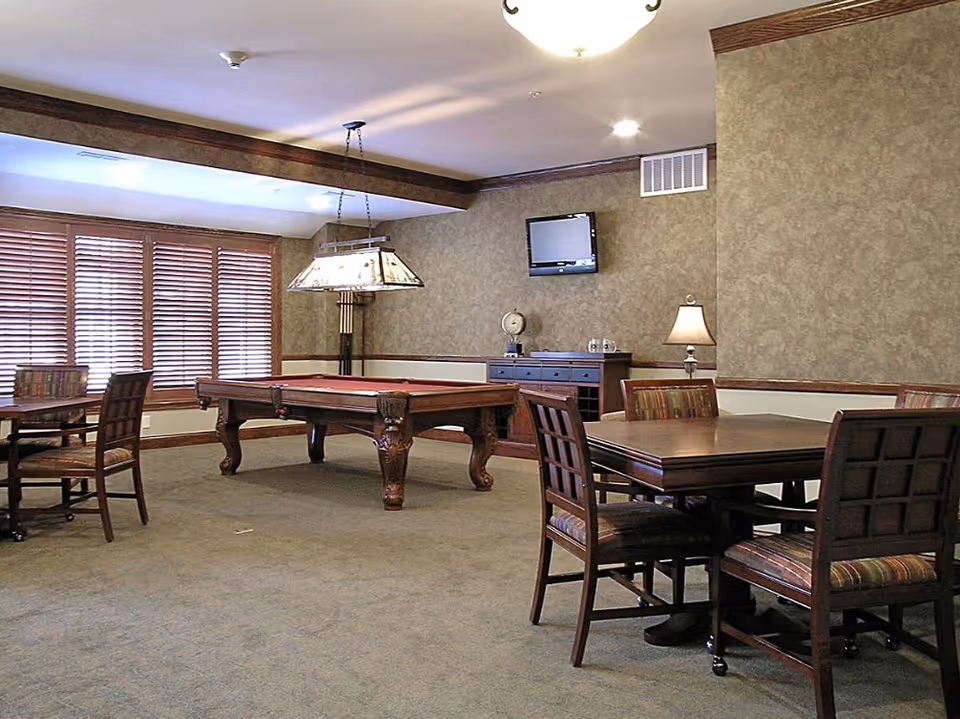 A spacious room with a pool table in the center, surrounded by wooden tables and chairs with patterned cushions. The room has beige walls with wood trim, a hanging light fixture above the pool table, a wall-mounted TV, and a sideboard with a lamp and decorative items. Large windows with wooden blinds allow natural light to enter.