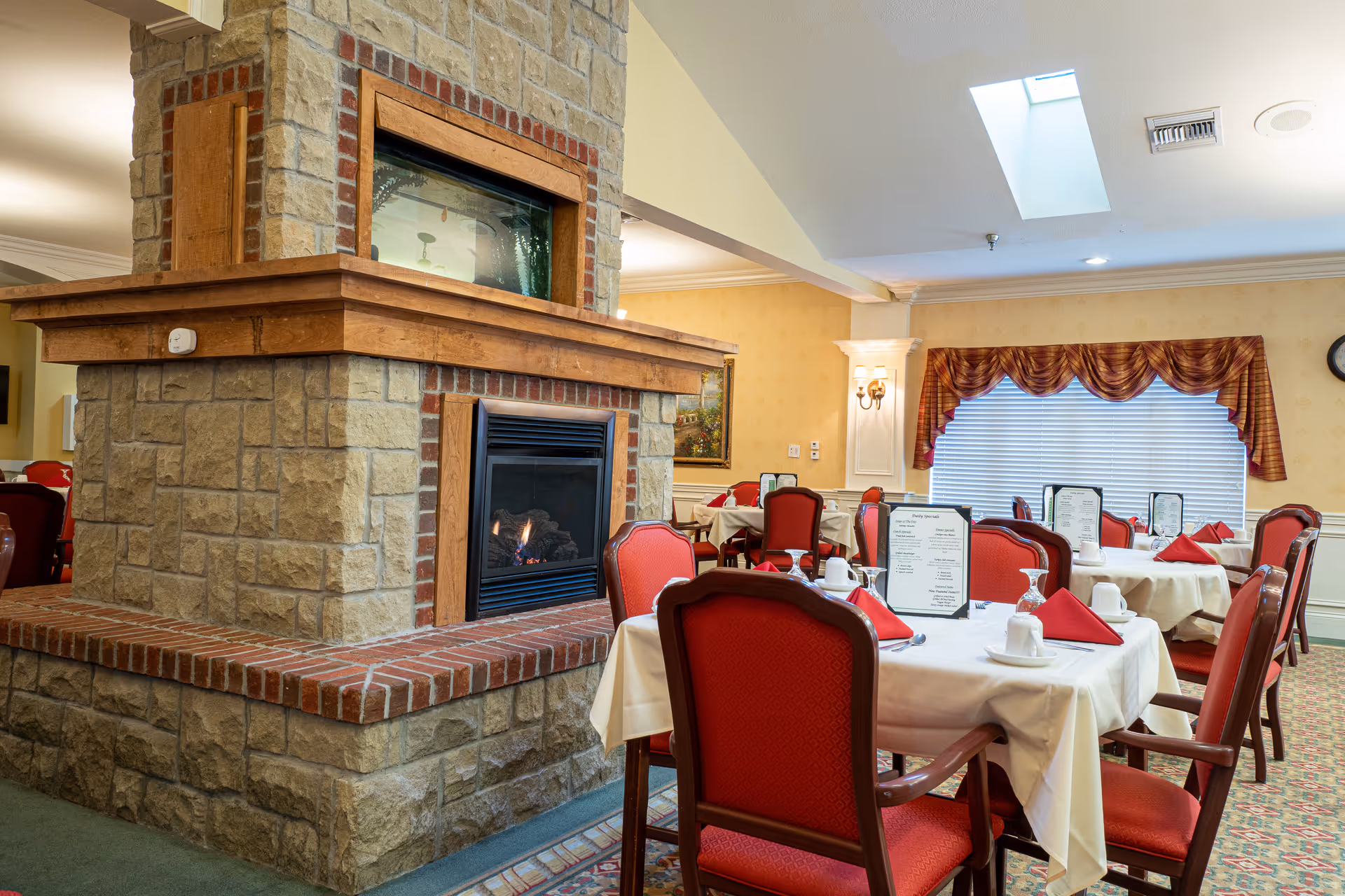 Dining room with a central stone fireplace and tables set with white tablecloths, red chairs, and red napkins.
