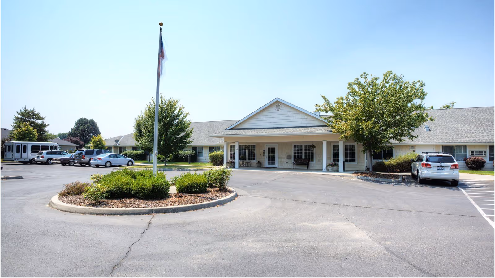 Single-story memory care building entrance with a circular driveway, flagpole, parked cars, and landscaping under a clear sky.