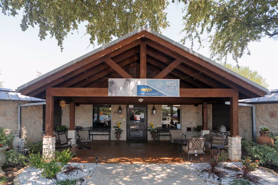Entrance of Lake Village Nursing & Rehabilitation Center featuring a covered porch with wooden beams, stone pillars, outdoor seating including chairs and benches, potted plants, and a sign above the door displaying awards and the facility name.