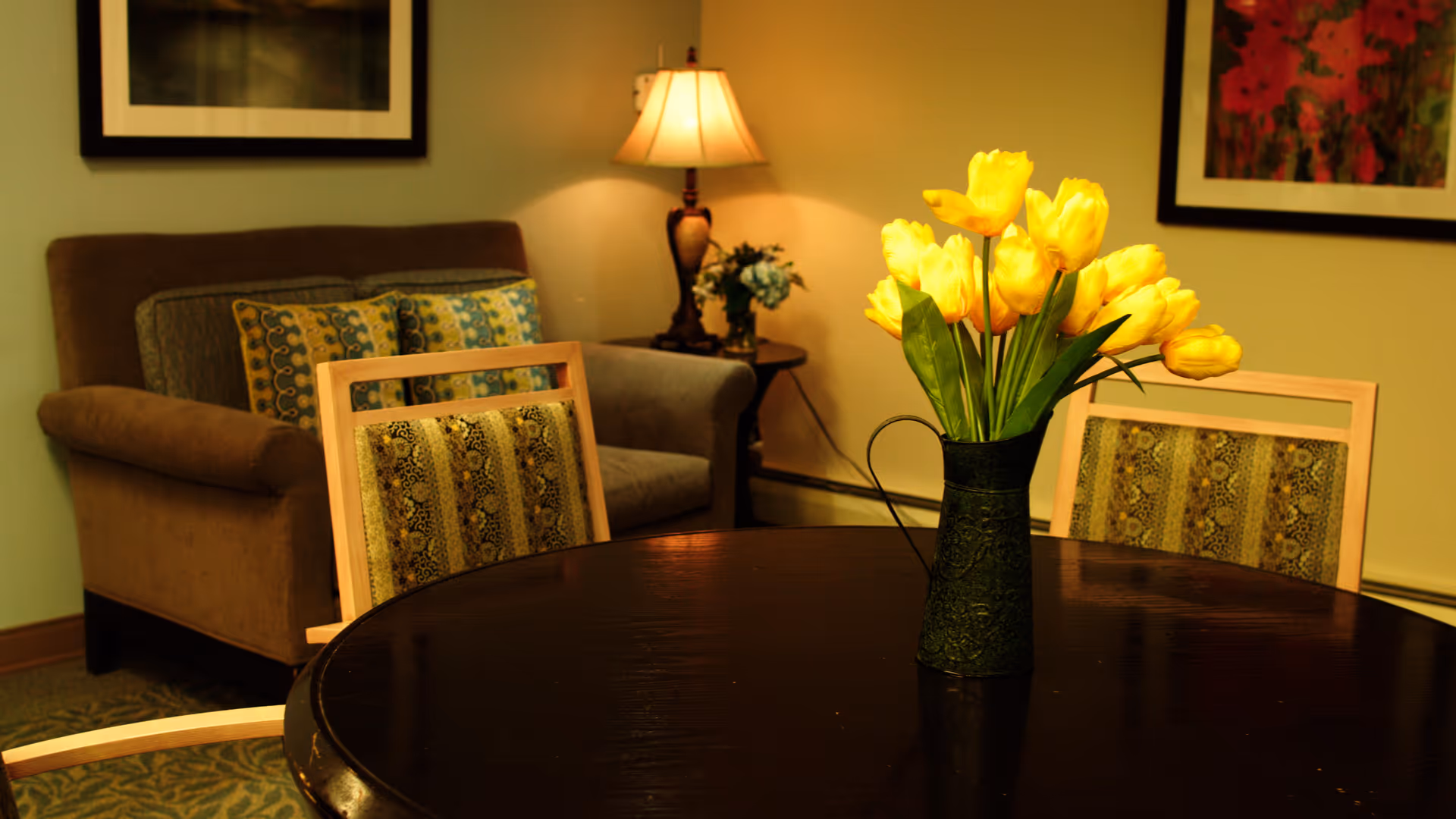 Round dark wood table with a vase of yellow tulips in a cozy seating area with chairs, a sofa and a lamp.