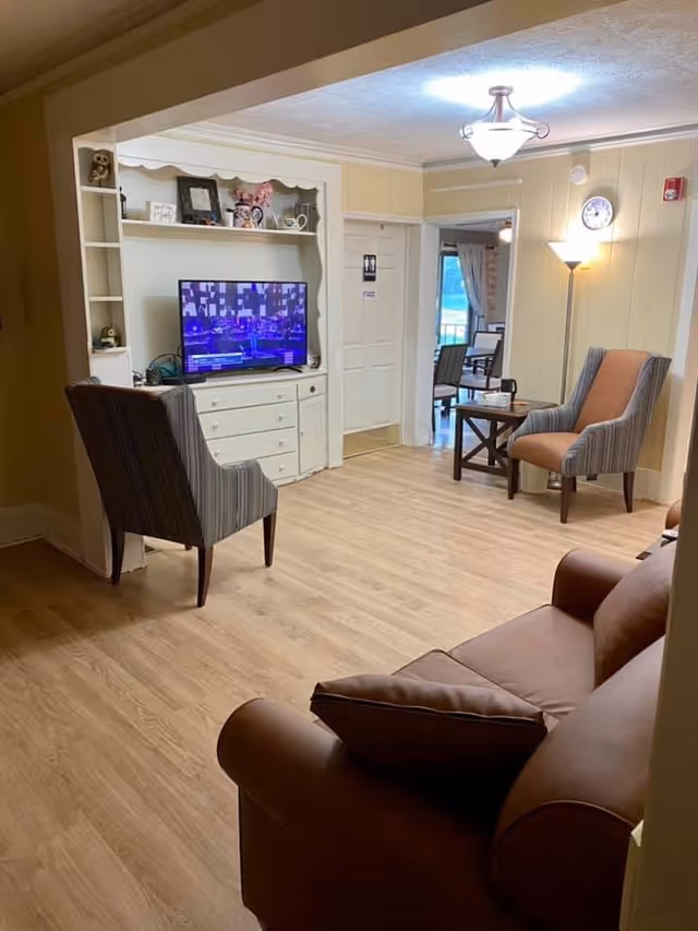 A cozy living room area with a brown leather sofa in the foreground, two striped armchairs, a small wooden side table, a floor lamp, and a flat-screen TV on a white cabinet with shelves displaying decorative items. The room has light wood flooring and cream-colored walls with a ceiling light fixture. A doorway leads to another room with additional seating and a window with curtains.