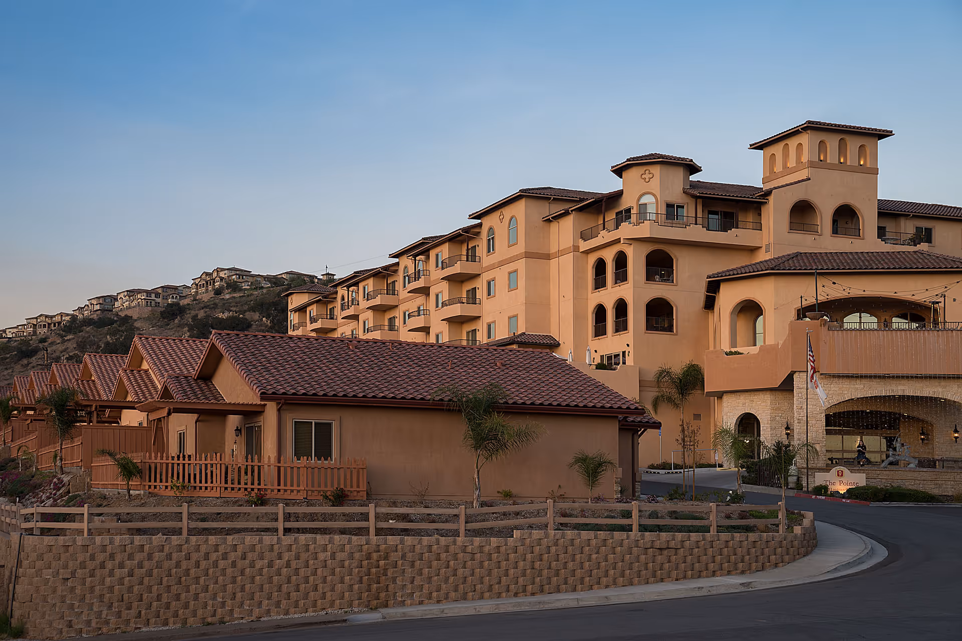 Exterior view of a senior living facility with multiple beige buildings featuring red-tiled roofs, balconies, and arched windows, set against a hillside with other houses in the background under a clear sky.