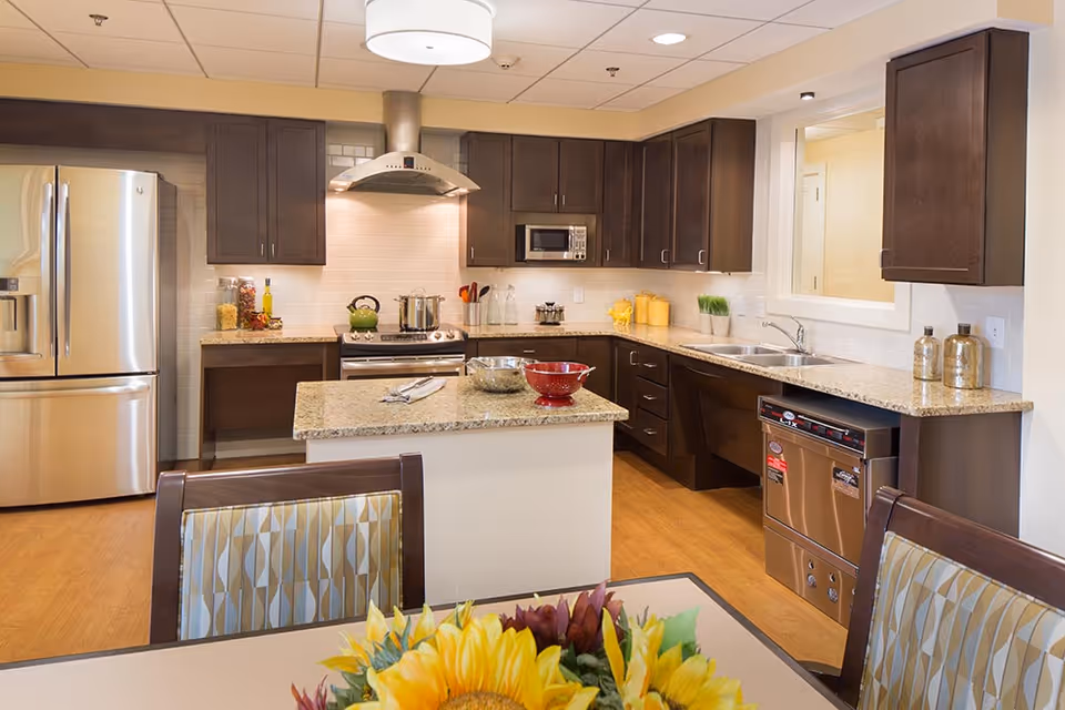 Modern kitchen in a senior living facility with dark wood cabinets, stainless steel refrigerator, stove with hood, microwave, dishwasher, and granite countertops. A small island with bowls is in the center, and a dining table with patterned chairs and a sunflower centerpiece is partially visible in the foreground.