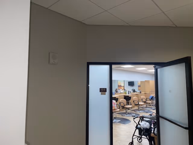 View through an open door labeled 108 into a room with multiple salon chairs and mirrors, resembling a hair salon or beauty care area. A person with a walker is partially visible inside the room.