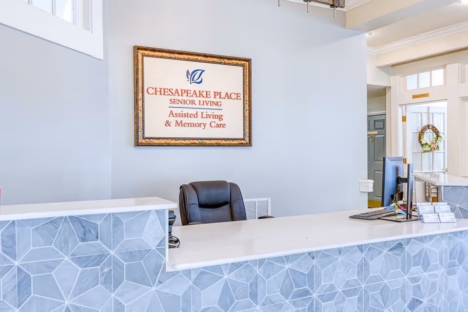 Reception desk area at Chesapeake Place Senior Living with a framed sign on the wall reading 'Chesapeake Place Senior Living Assisted Living & Memory Care'. The desk has a geometric patterned front and a computer monitor on top, with a black office chair behind it.