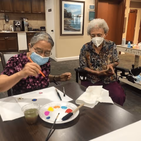 Two elderly women wearing face masks are seated at a table engaged in a painting activity. One woman is actively painting on a piece of paper with a brush, while the other watches. The table has a palette with various paint colors, brushes, a cup with green liquid, and some tissues. The background shows a kitchen area and a wall with a framed picture.