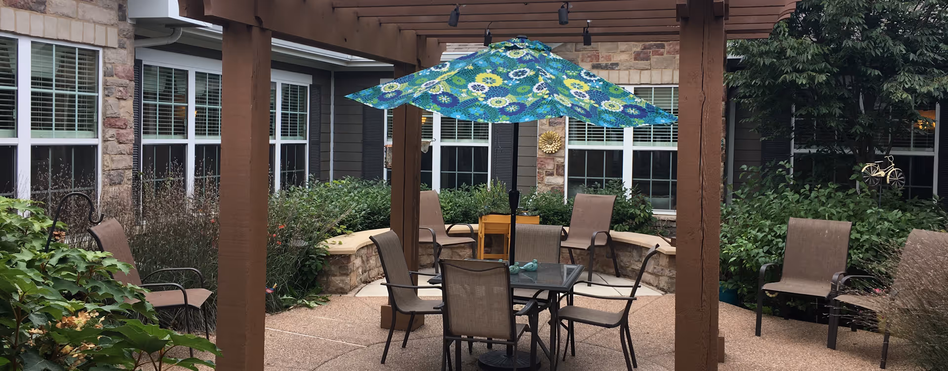 Outdoor patio area with a wooden pergola, a glass table with four chairs, a colorful floral umbrella, additional chairs around the perimeter, and surrounding greenery and shrubs in front of a building with stone and siding exterior and multiple windows.