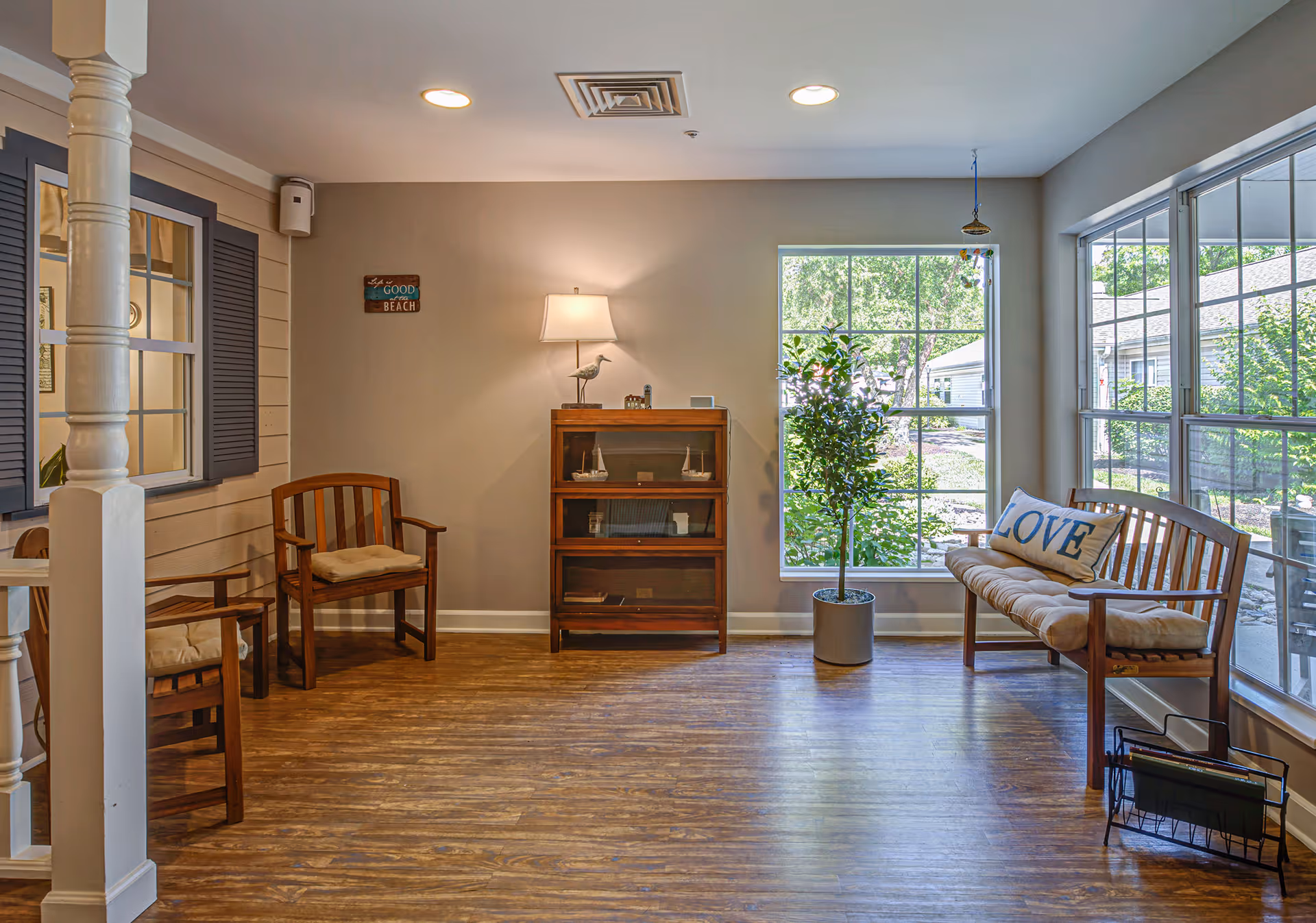 A cozy sitting area with wooden chairs and a bench with a cushion and a pillow that says 'LOVE'. There is a small wooden cabinet with a lamp and decorative items on top, a potted plant near a large window, and a sign on the wall that reads 'Life is good at the BEACH'. The room has wood flooring and large windows letting in natural light.