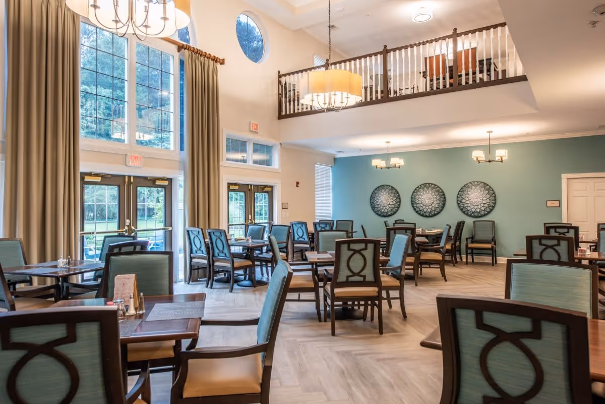 A spacious dining room with multiple wooden tables and chairs featuring light blue upholstery. Large windows with beige curtains allow natural light to fill the room. The walls are painted light blue and decorated with three circular wall art pieces. There are two chandeliers hanging from the ceiling, and a balcony area overlooks the dining space.