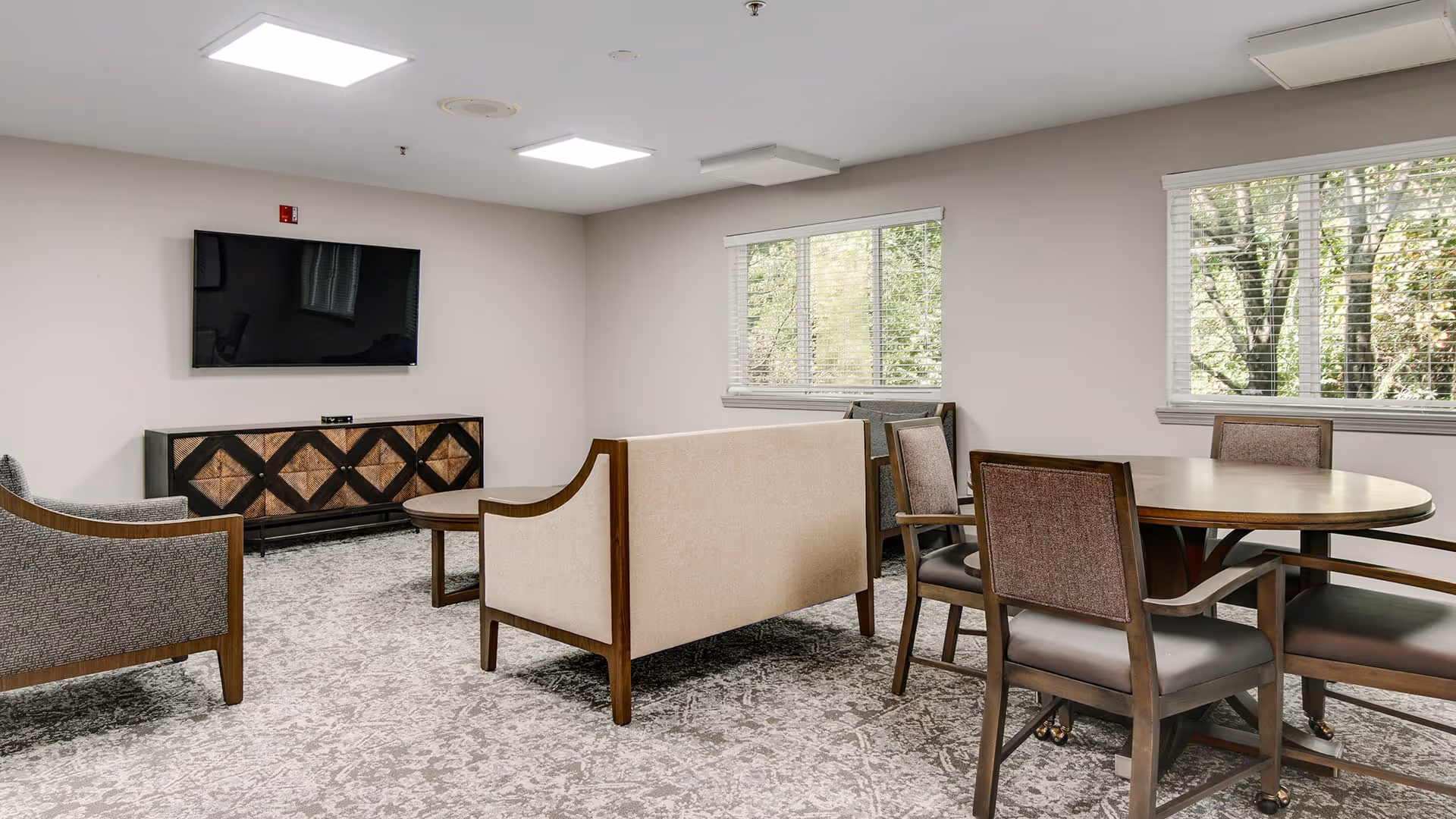 A well-lit living room area with a mounted flat-screen TV on the wall above a wooden cabinet with a geometric pattern. The room features a beige sofa, two armchairs, a round wooden table with four chairs, and two large windows with white blinds showing greenery outside. The floor is covered with a patterned carpet.