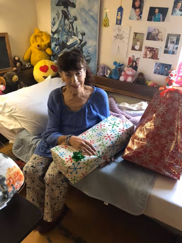 An elderly woman sitting on a bed in a room decorated with stuffed animals, photos, and drawings on the wall. She is holding a wrapped gift box with colorful snowflake patterns and a green bow. There is another large wrapped gift next to her on the bed.