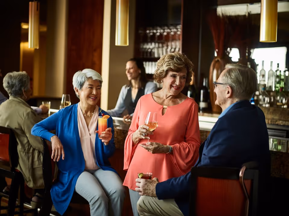 Three elderly people socializing and enjoying drinks at a bar inside a senior living facility. Two women and one man are smiling and conversing, with a bartender in the background and shelves of glasses and bottles behind the bar.
