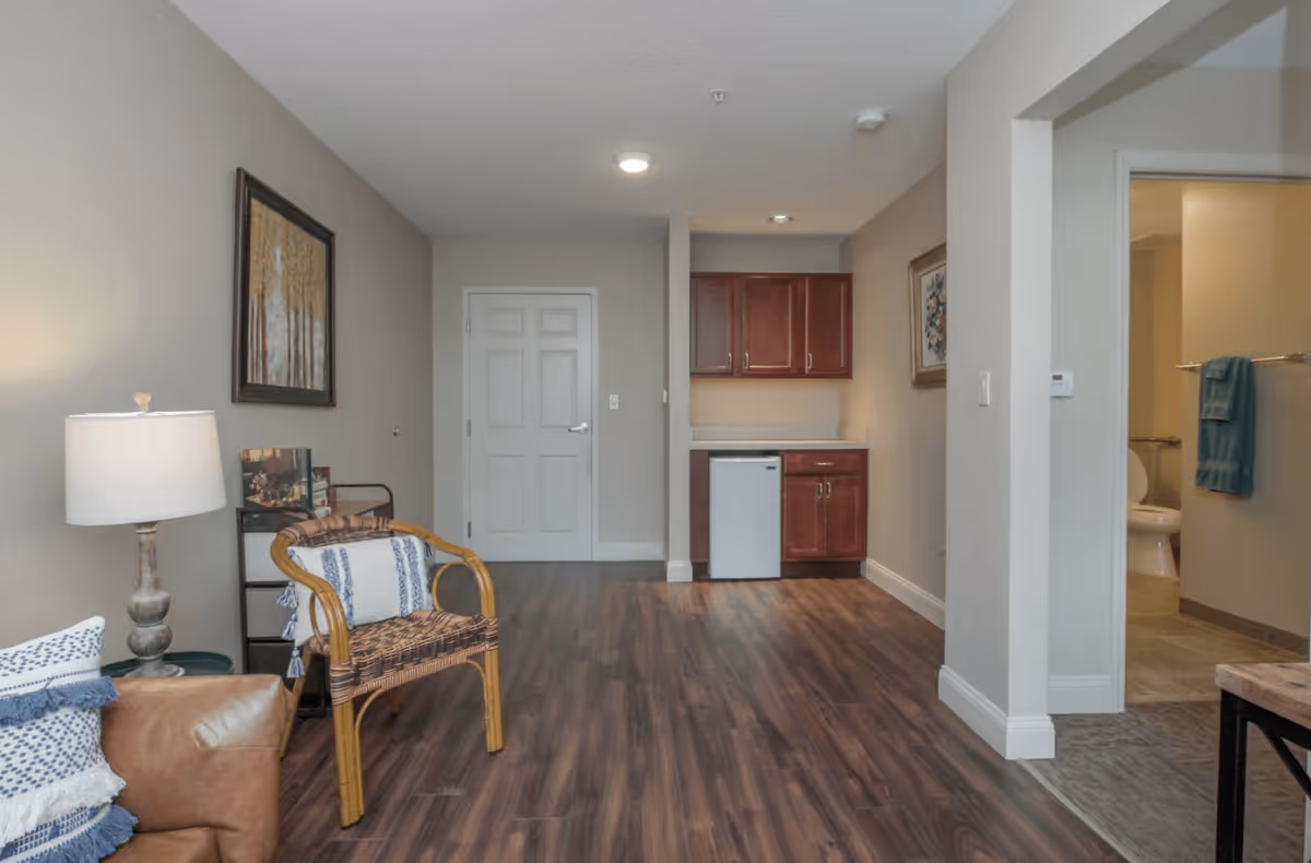 Interior view of a senior living facility room with wood flooring, a small kitchenette with cabinets and a mini fridge, a wicker chair with a cushion, a leather couch with pillows, a floor lamp, and a bathroom visible through an open doorway.