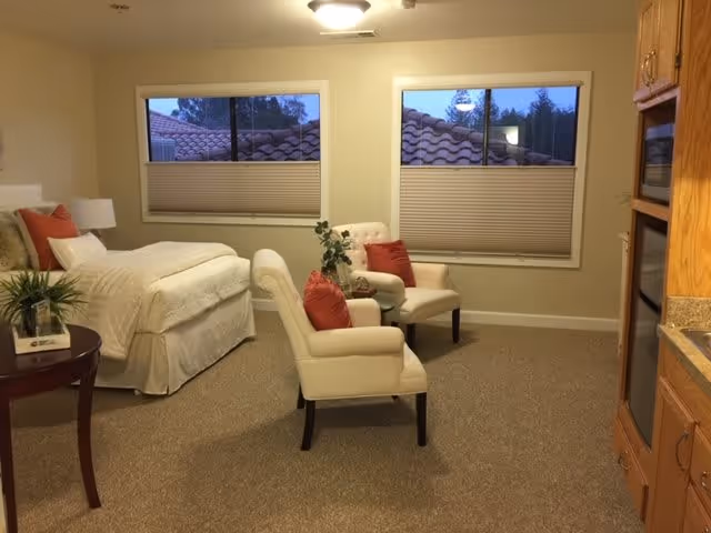 A cozy bedroom in an assisted living facility featuring a bed with white bedding and orange accent pillows, two beige armchairs with orange pillows, a small round table with a plant, two windows with blinds partially closed, and a kitchenette area with wooden cabinets and appliances.