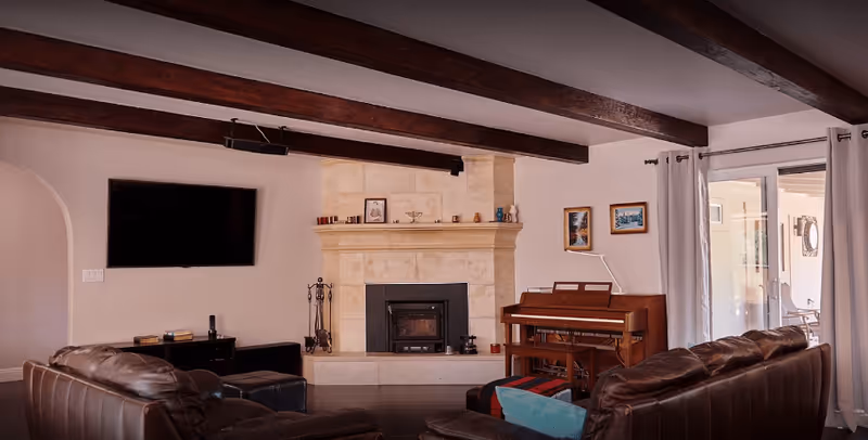 A cozy living room with two brown leather sofas facing a stone fireplace. Above the fireplace are decorative items and framed pictures. A wooden piano is placed against the wall next to a large window with curtains. A flat-screen TV is mounted on the wall opposite the sofas. The ceiling features exposed wooden beams.