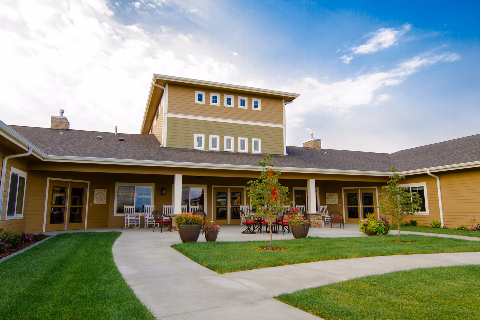 Exterior view of a senior living facility with a covered patio area featuring several rocking chairs and tables with chairs. The building has a multi-level roof and is surrounded by a well-maintained lawn, small trees, and flower pots. The sky is partly cloudy with blue patches.