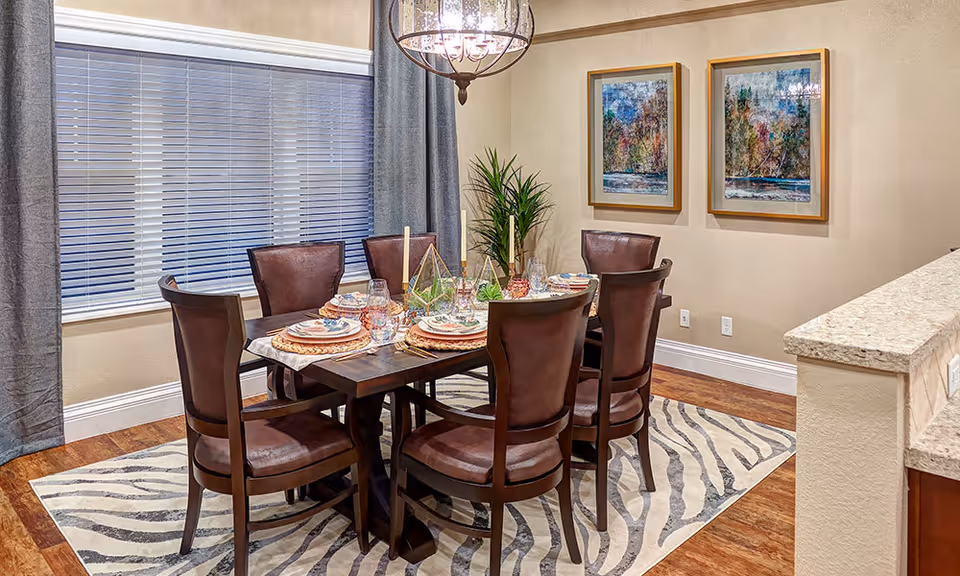 A dining room with a dark wooden table set for six people with plates, glasses, and candles. The chairs are brown leather with wooden frames. The room has a large window with closed blinds and gray curtains. Two framed paintings hang on the beige wall, and a green plant is placed in the corner. The floor has a wood finish with a zebra-patterned rug under the table. A chandelier with multiple lights hangs above the table.