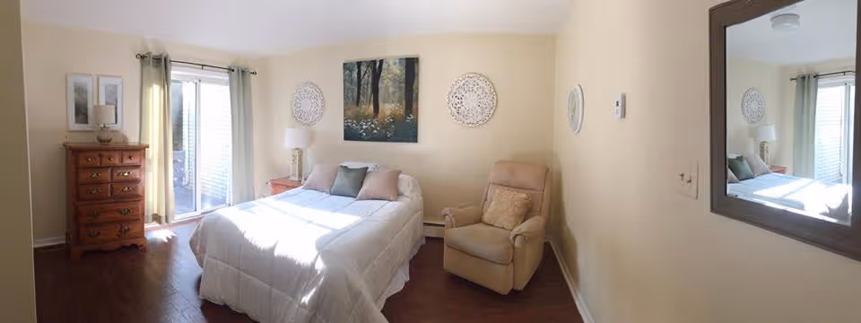 Sunlit bedroom with a bed, armchair, wooden dresser, sliding glass door with curtains, and wall decor.