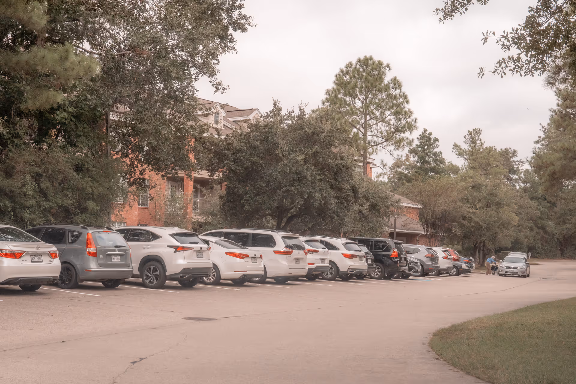 Parking lot with multiple cars parked in a row next to a senior living facility building partially visible behind trees and greenery. An elderly person with a walker is seen near a silver car at the far end of the parking lot.