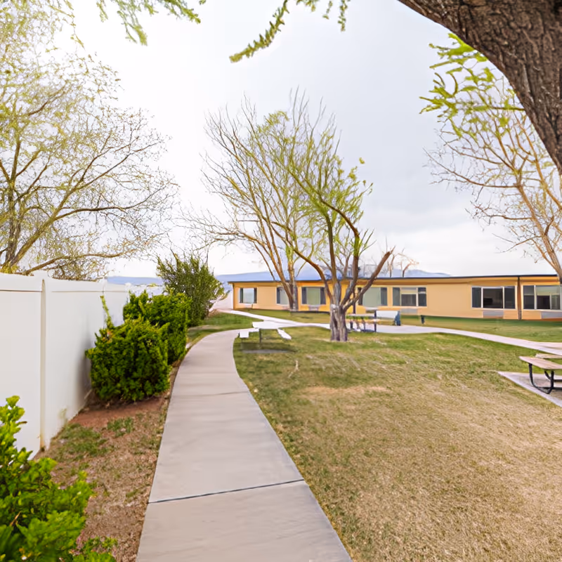 A paved walkway curves through a grassy outdoor area with leafless and partially leafed trees. There are bushes along a white fence on the left side and picnic tables on the right. In the background, there is a single-story building with multiple windows under a cloudy sky.