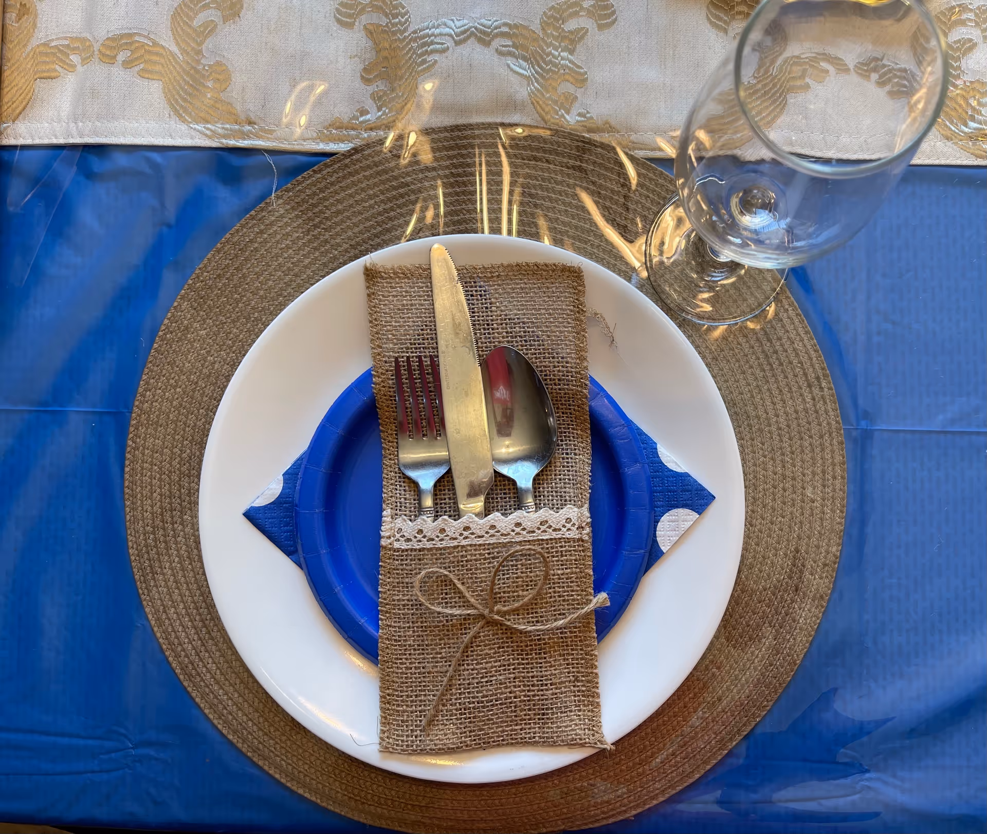 Top view of a dining table setting with a white plate, a smaller blue plate, a blue and white napkin, and silverware (fork, knife, spoon) wrapped in a burlap holder tied with a string bow. A clear wine glass is placed to the right of the plates on a brown woven placemat over a blue tablecloth.