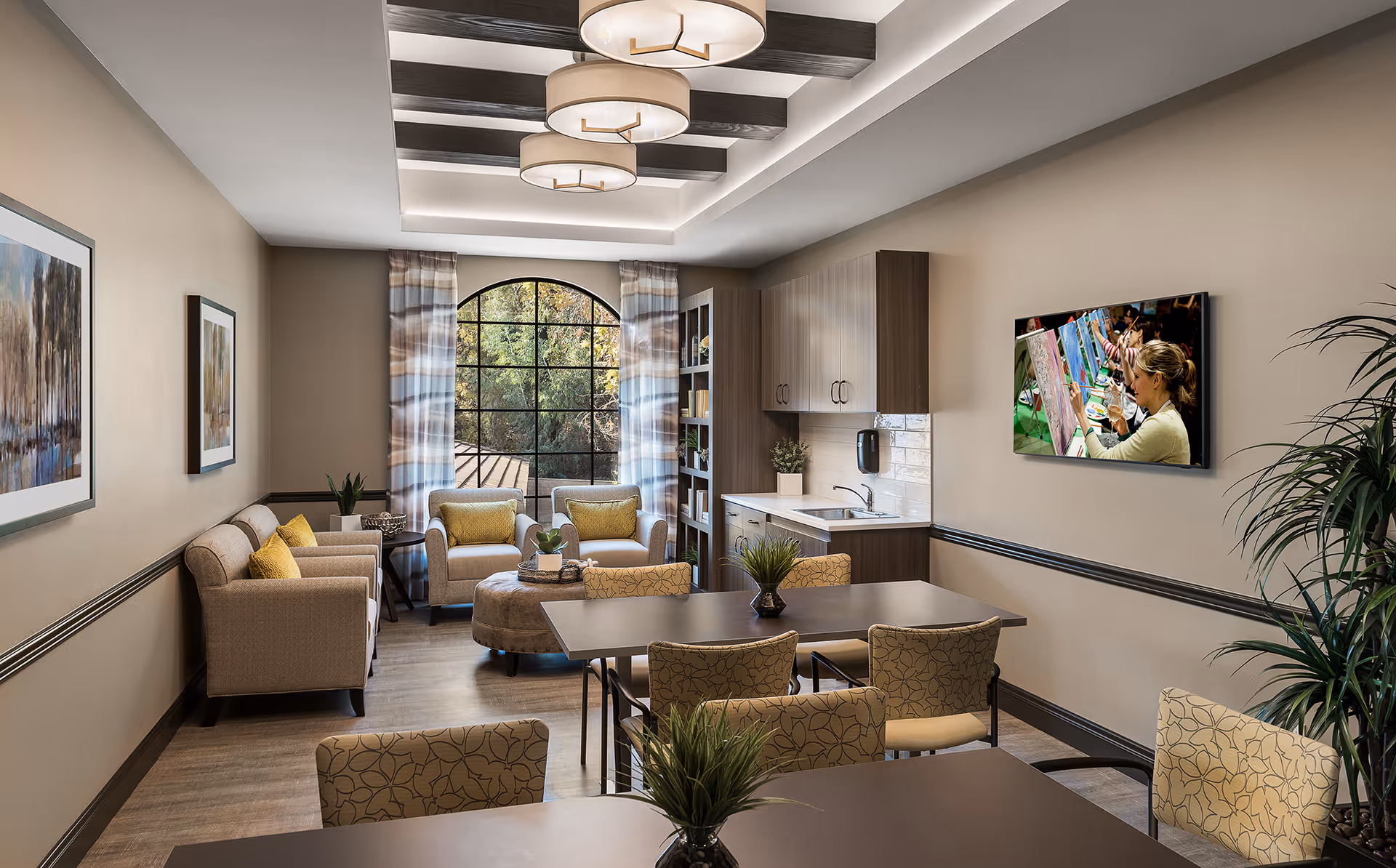 A well-lit common room with beige walls and wood flooring featuring a seating area with armchairs and a round ottoman near a large arched window with patterned curtains. There are two tables with patterned chairs and small potted plants on them. A kitchenette with cabinets, a sink, and a soap dispenser is on one side, and a flat-screen TV mounted on the wall displays people painting. The ceiling has decorative wooden beams and three round pendant lights.