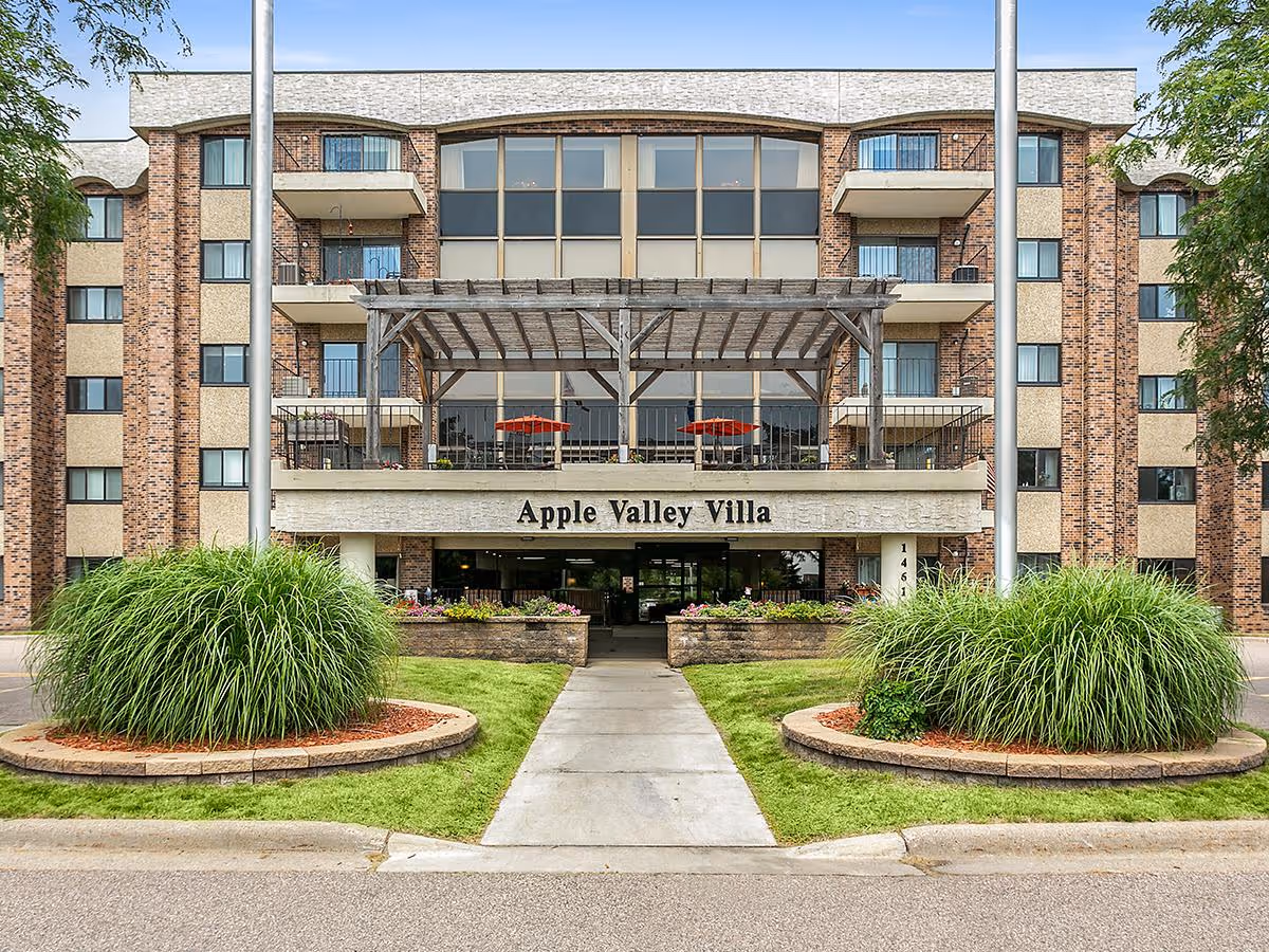 Front exterior view of a multi-story brick and stucco building with balconies, a covered patio area with red umbrellas, and a sign that reads 'Apple Valley Villa'. The entrance is flanked by landscaped greenery and a sidewalk leading up to the door.