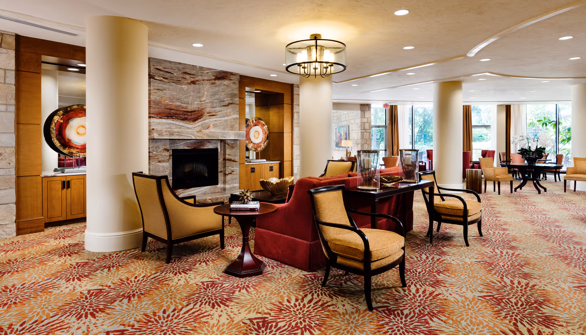 A spacious and well-lit senior living facility common area with patterned carpet, multiple seating arrangements including armchairs and sofas, a decorative fireplace with stone surround, large windows letting in natural light, and decorative elements such as vases and artwork.