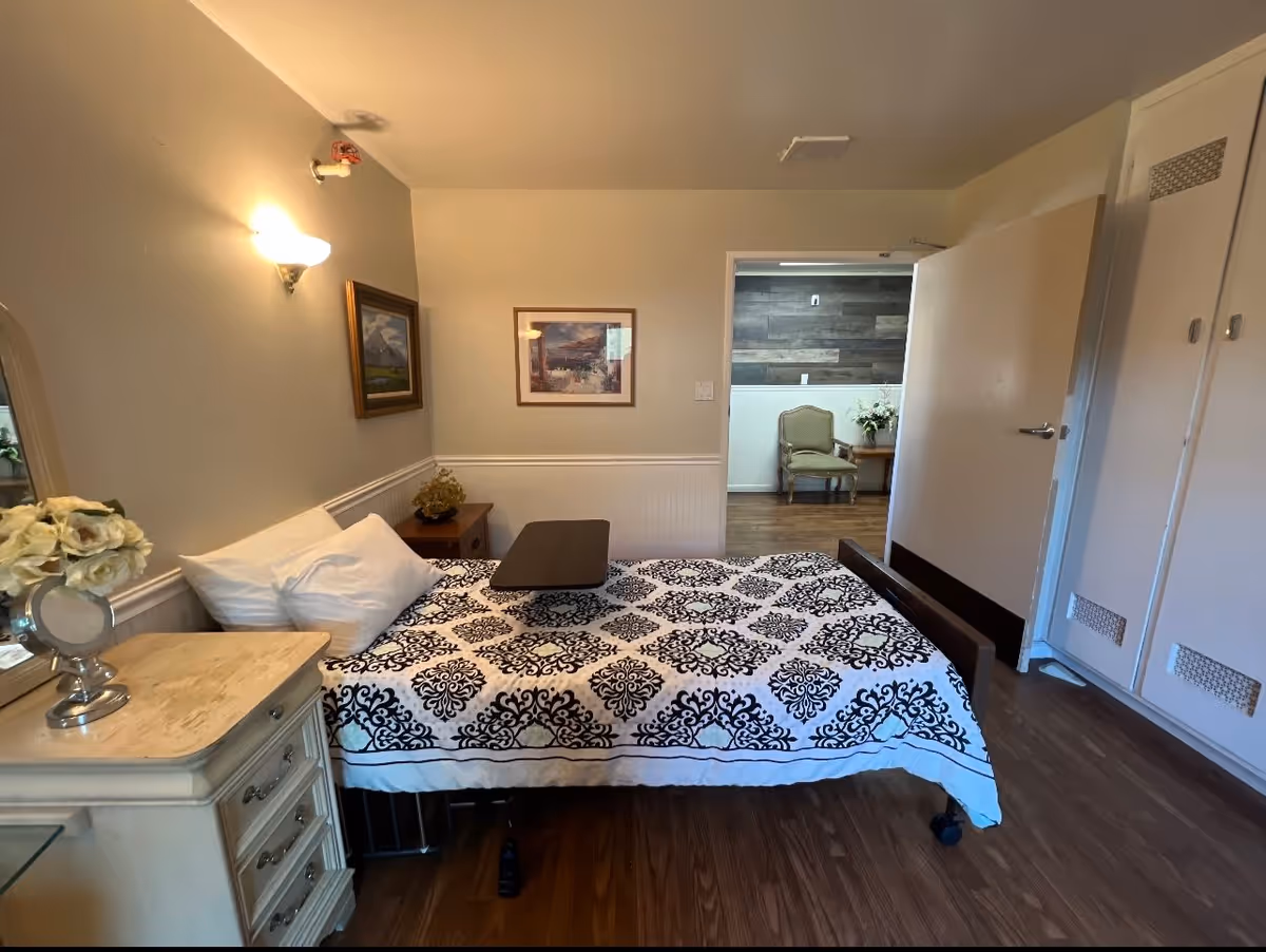 A bedroom in a senior living facility with a bed covered in a black and white patterned bedspread, two pillows, and a small overbed table. There is a cream-colored dresser with a vase of white flowers and a mirror on the left side. The walls are light-colored with two framed pictures hanging. The door is open, revealing a hallway with a chair and a small table with flowers. The floor is wooden.
