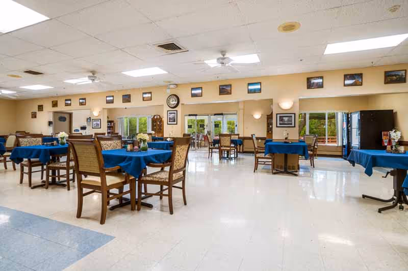 A spacious dining room with several tables covered in blue tablecloths and surrounded by wooden chairs with cushioned seats. The room has large windows letting in natural light, a clock on the wall, and framed pictures above the windows. The floor is tiled, and the ceiling has recessed lighting and ceiling fans.