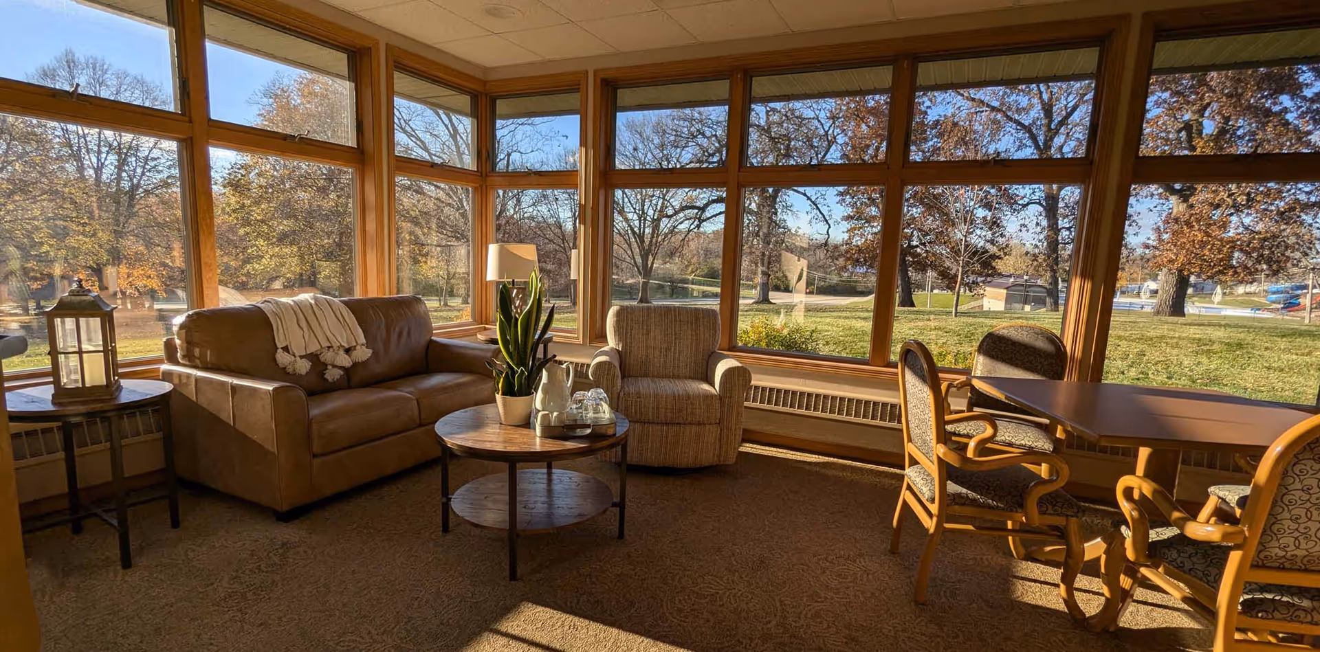 Sunlit common room with a sofa, armchair, coffee table and dining table in front of large panoramic windows overlooking a grassy outdoor area.