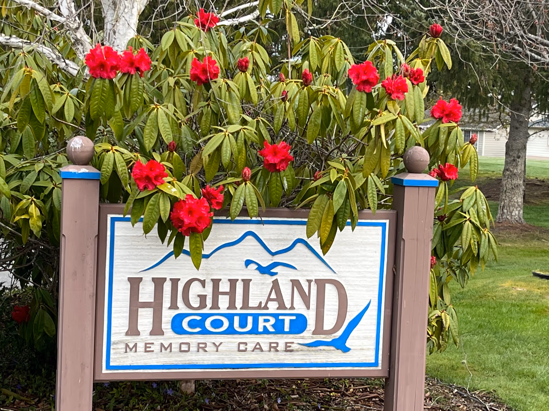 A wooden sign for Highland Court Memory Care surrounded by green bushes with bright red flowers. The sign features blue and brown text with a mountain and bird design. The background includes grass and trees.
