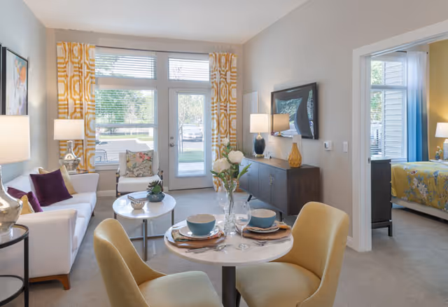 A bright and cozy senior living apartment featuring a small round dining table set for two with bowls, plates, and wine glasses. The living area includes a white sofa with purple pillows, a small round coffee table, and a chair with a floral cushion. Large windows with yellow patterned curtains let in natural light, and a door leads outside. A dark wood sideboard with lamps and decorative items is against the wall, and a bedroom with a yellow bedspread is visible through an open doorway.