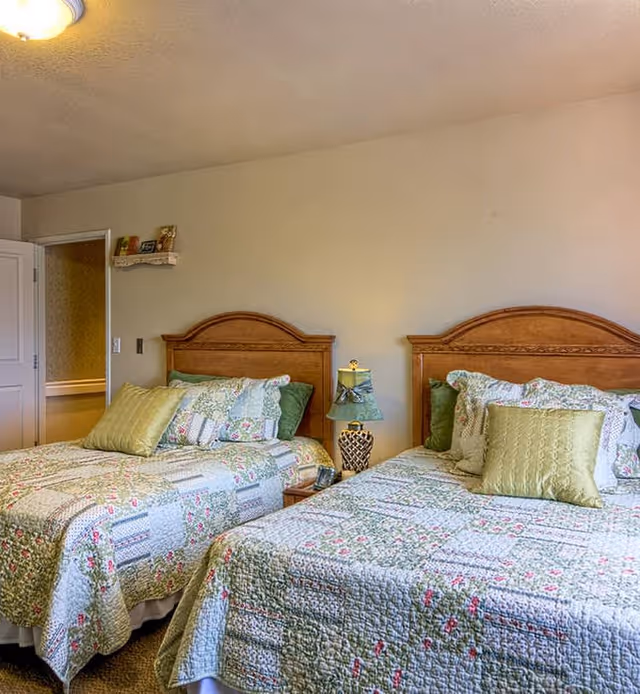 A bedroom with two twin beds featuring wooden headboards and floral patterned quilts in shades of green, white, and pink. Each bed has multiple pillows, including green accent pillows. A small nightstand with a decorative lamp is positioned between the beds. The room has beige walls and a door leading to another area.