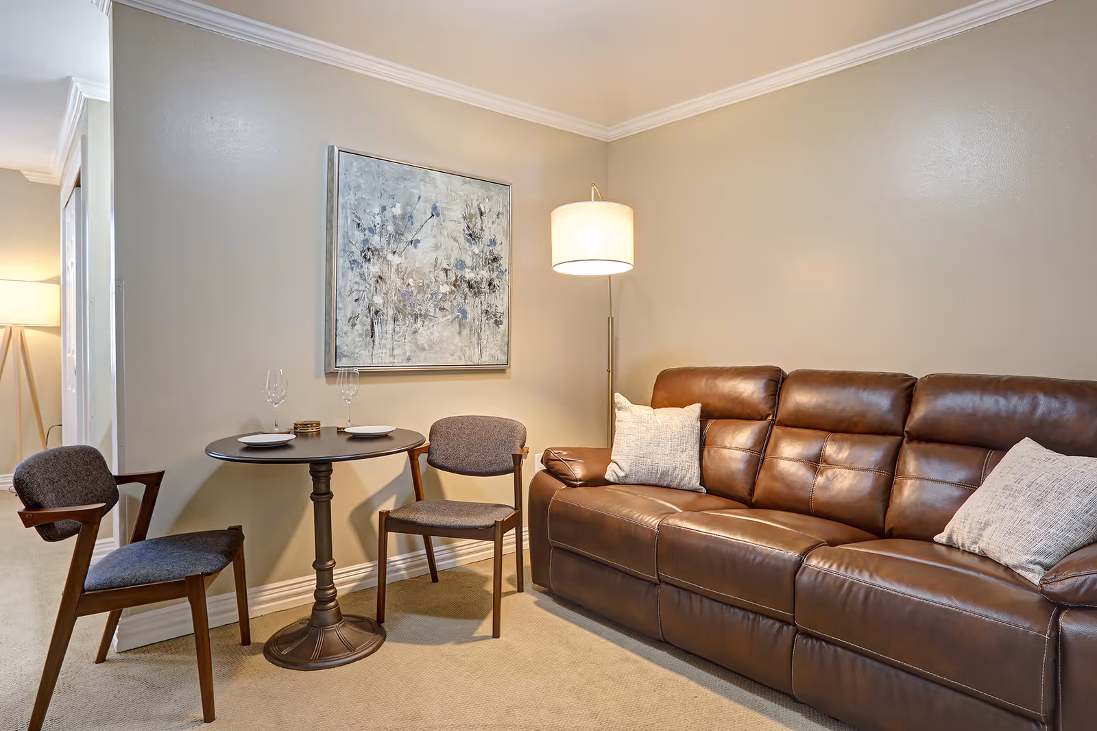 A cozy living area featuring a brown leather sofa with two light-colored pillows, a small round table set with two plates and wine glasses, and two wooden chairs with gray cushions. A floor lamp with a white shade stands behind the sofa, and a large abstract painting hangs on the beige wall.