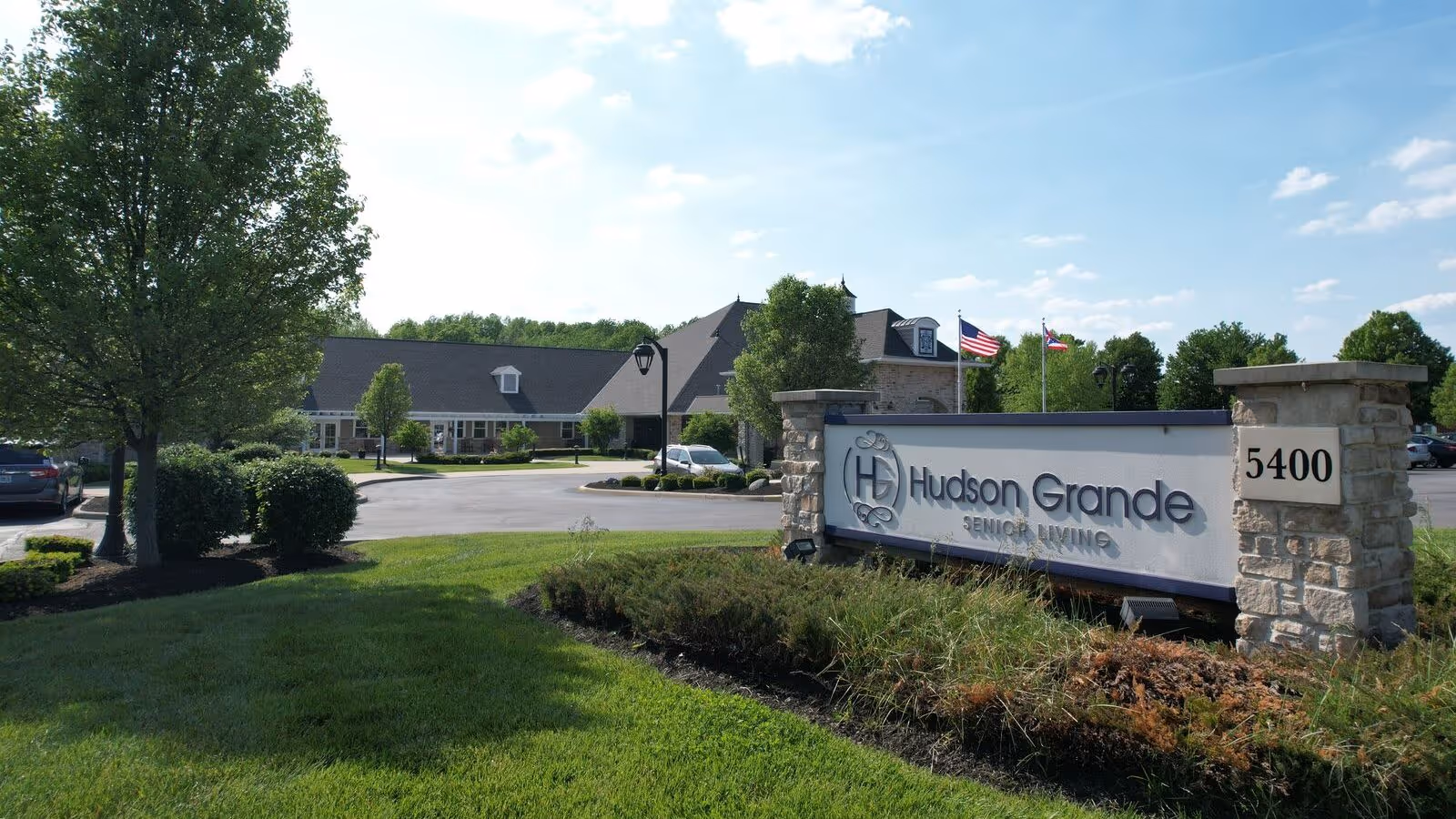 Exterior view of Hudson Grande Senior Living facility with a large sign displaying the name and address 5400. The building is surrounded by green grass, trees, and a parking lot with cars. Two flags are visible near the building under a partly cloudy sky.