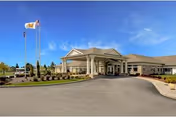 Front exterior view of a single-story senior living facility building with a covered entrance, two flagpoles with flags, landscaped grounds, and a clear blue sky.