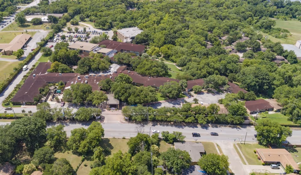 Aerial view of The Villages of Dallas senior living facility surrounded by dense green trees and vegetation. The complex consists of multiple connected buildings with brown roofs, parking lots, and nearby roads with a few cars. The area around the facility is lush and green with residential houses and open spaces.